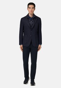 Námornícka modrá sako s textúrovanou tkaninou, s dvoma prednými vreckami, vrstvené cez tmavú páperovú bundu, kombinované s námorníckymi nohavicami a čiernymi teniskami.