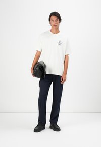 Junger Mann, der ein weißes bedrucktes T-Shirt, eine marineblaue Hose und schwarze Schuhe trägt und in seiner linken Hand eine gefaltete schwarze Lederjacke hält.