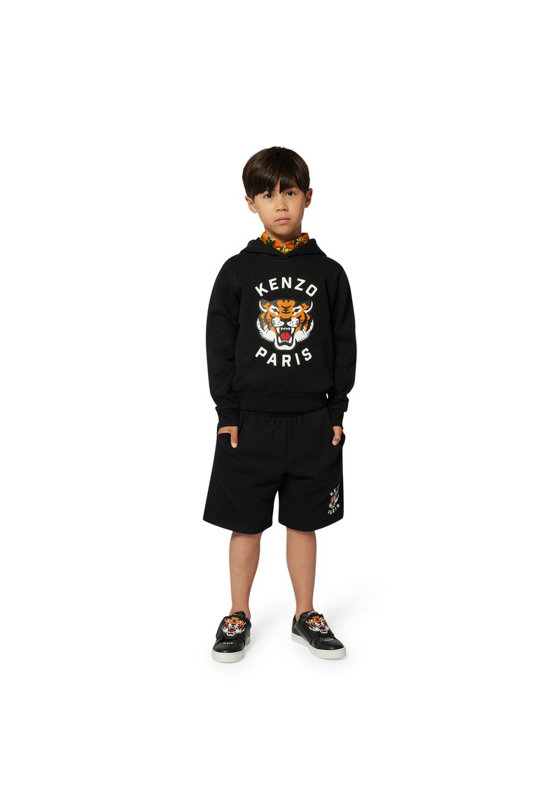 Zwarte hoodie met tijgerafbeelding, gecombineerd met zwarte shorts. Zwarte schoenen met tijgerdetail. Kleurrijke kraag zichtbaar eronder.