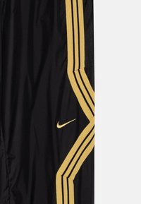 Svarte sportbukser med gule og svarte stripete sidepaneler og en liten gull Nike-sjekk logo. Laget av lett, teksturert stoff.