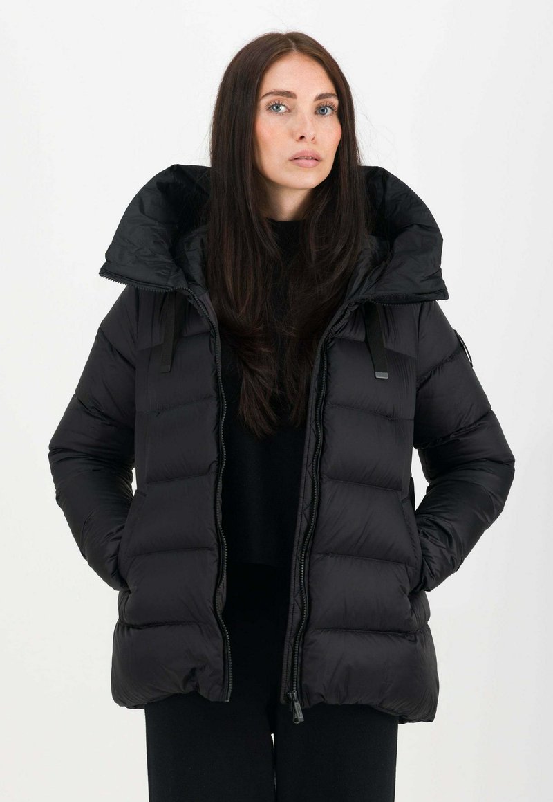 Jeune femme aux longs cheveux bruns portant une veste matelassée noire avec un col montant et les mains dans les poches, face à l'avant sur fond blanc.