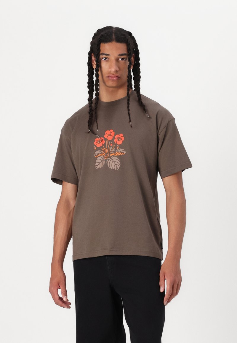 Brun bomull T-skjorte med et blomsterdesign i oransje og grått. Korte ermer, rund hals og ledig passform. Sorte bukser nedenfor.