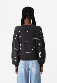 Donkergrijze sweater met oversized mouwen, voorzien van witte borduring in een hemels patroon, geribbelde manchetten en een ronde halslijn. Gecombineerd met lichtblauwe jeans.