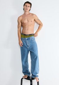 Jeans in denim blu con una vestibilità ampia e una texture scolorita, abbinati a una cintura nera e verde, in piedi su uno sgabello nero contro un muro bianco.