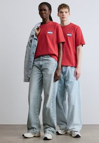 T-shirts vermelhas de algodão com um pequeno gráfico azul e branco, combinadas com jeans azuis claros de cintura alta e ténis brancos. Casaco de ganga usado.