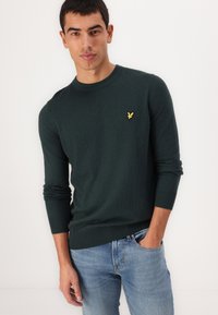 Dark green knitted sweater with a crew neckline and long sleeves, featuring a small yellow bird logo on the left chest. Worn with blue jeans.