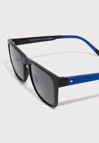 Ochelari de soare negri cu lentile gri, având un braț texturat albastru și un mic accent cu logo pe templu. Design dreptunghiular elegant și modern.