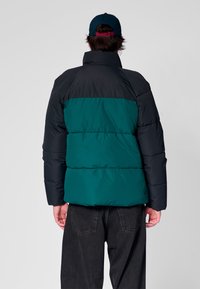 Kurtka puchowa z czarnymi i turkusowymi panelami, z wysokim kołnierzem i pikowanym wzorem. W połączeniu z ciemnymi dżinsami i granatową czapką.