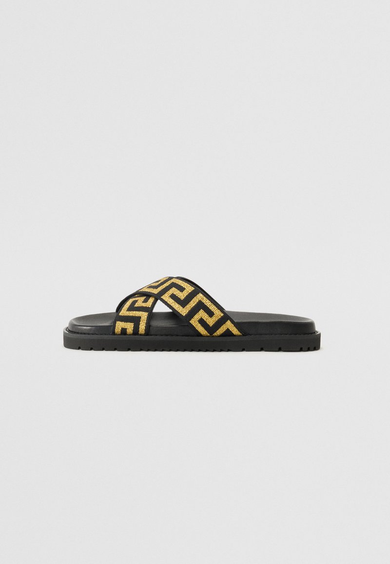 Black slide sandal with a textured black sole. Features thick straps adorned with a gold geometric pattern. Flat design, suitable for casual wear.