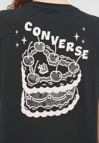 Μαύρο πουκάμισο με γραφικό σχέδιο ενός πολύστιχου κέικ με κεράσια στην κορυφή και τη λέξη "CONVERSE" από πάνω, όλα σε λευκό.