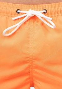 Costume da bagno arancione con una texture liscia e leggera. Presenta un cordino bianco passato attraverso occhielli neri in vita.