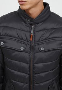 Doudoune noire matelassée avec un col montant, un motif matelassé horizontal, une fermeture éclair à l'avant et deux poches poitrine à boutons-pression.