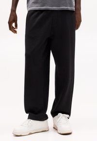 Zwarte katoenen joggingsbroeken hebben een losse pasvorm, elastische tailleband en zijzakken, gecombineerd met witte sneakers en een grijs shirt.
