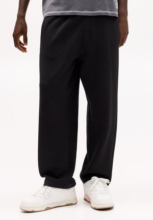I pantaloni sportivi neri in cotone hanno una vestibilità ampia, una cintura elastica e tasche laterali, abbinati a sneaker bianche e una maglietta grigia.