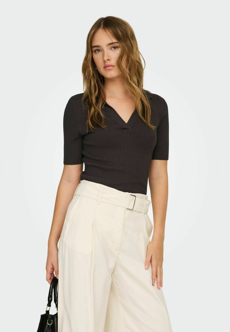 Femme portant un haut noir ajusté à col en V avec des manches jusqu'au coude et un pantalon large crème taille haute, tenant un petit sac à main noir.