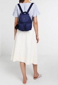 Sac à dos en tissu bleu foncé avec bretelles réglables, deux poches avant et une finition texturée. Il est orné d'un pompon noir.