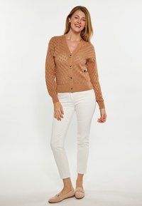 Cardigan lavorato a maglia color marrone chiaro con un motivo a diamante, scollo a V e chiusura con bottoni, abbinato a pantaloni bianchi aderenti e mocassini beige.