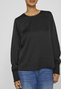 Maglia nera a maniche lunghe con una texture liscia, collo rotondo e polsini arricciati; abbinata a jeans in denim blu.
