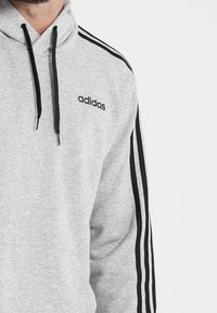 Felpa grigia realizzata in tessuto misto cotone, con strisce nere sulle maniche e un logo frontale. Include un cappuccio regolabile con cordini e polsini a coste.
