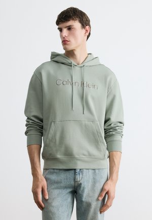 Νέος άνδρας που φοράει ανοιχτό πράσινο φούτερ Calvin Klein με τσέπη μπροστά και ανοιχτό μπλε τζιν, κοιτάζοντας προς τα πλάγια σε απλό φόντο.