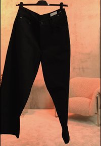 Μαύρο τζιν με ψηλή μέση κρεμασμένο σε κρεμάστρα σε ένα απαλά φωτισμένο δωμάτιο με μια μπεζ υφασμάτινη πολυθρόνα στο φόντο.