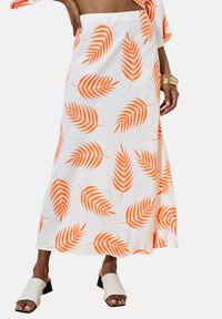 Jupe longue blanche avec imprimé de feuille de palmier orange, présentant un design fluide et un tissu léger. Associée à des sandales à talons carrés écrues.