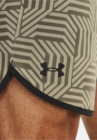Vzorované sportovní šortky v šedé a krémové barvě, s geometrickým designem, černým lemováním a logem Under Armour na dolním levém rohu.