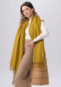 Sciarpa gialla senape, tessuto morbido, con bordi frangiati, indossata sopra una maglietta bianca aderente, abbinata a pantaloni beige a vita alta, su uno sfondo neutro.