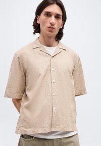 Νεαρός άνδρας με σκούρα μαλλιά μέχρι τους ώμους, φορώντας μπεζ κοντομάνικο πουκάμισο με κουμπιά πάνω από λευκό t-shirt, σε απλό φόντο.