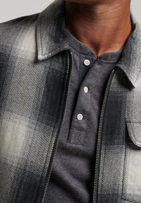 Kariertes Hemdjacke mit schwarz-weißem Karomuster, darunter ein graues Henley, graue Knöpfe und ein Kragen mit Reißverschluss.