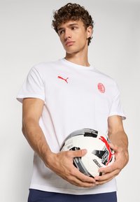 Hvid bomulds T-shirt med rødt Puma-logo og AC Milan-emblem, båret af en mandlig model, der holder en sort og hvid fodbold.