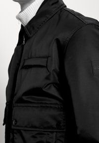 Svart nylonjacka med hög krage, frontdragkedja och två bröstfickor. Har en slät yta och ett märkespatch på ärmen.