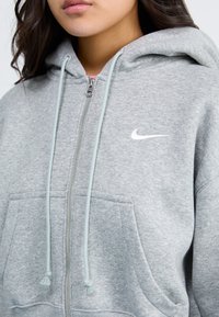 Harmaa vetoketjuhuppari pehmeästä kankaasta, jossa on kengurupocket, pastellinsiniset nyörit ja valkoinen Nike-logo rinnassa.