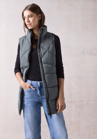 Dunkelgrüne, gesteppte Weste mit hohem Kragen und Reißverschluss, kombiniert mit hellblauen, lässig geschnittenen Jeans. Minimalistisches Design und glatte Textur.