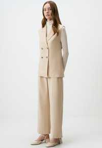 Beige op maat gemaakt vest met dubbelgebreide knopen, gecombineerd met losvallende beige broek en lichtgekleurde blokhak schoenen.