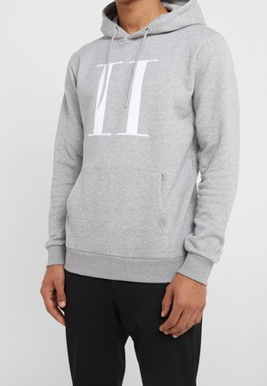 Mężczyzna w jasno szarej bluzie z kapturem z białym nadrukiem "VII", czarnych spodniach, z rękami swobodnie opuszczonymi wzdłuż ciała na tle jednolitego jasnego tła.