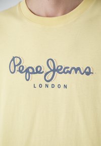 Geltonas medvilninis marškinėliai su ryškiu mėlynu grafiniu logotipu, kuriame rašoma "Pepe Jeans LONDON", su banguota dryžuota struktūra.