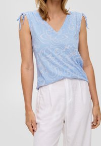 Donna che indossa un top senza maniche di colore azzurro chiaro con un motivo floreale e dettagli a cravatta sulle spalle, abbinato a pantaloni bianchi larghi a vita alta.