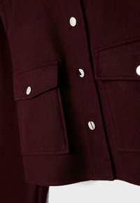 Burgundy wool blend jacket featuring silver snap buttons and two front pockets with flap closures. Smooth texture with a tailored design.