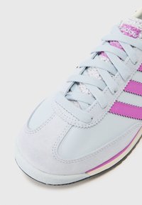 Ljusblå sneaker med mocka och syntetiska material. Har rosa ränder, strukturerade snören och en rund tå. Gummisulor med färgaccenter.