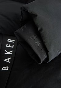Primo piano della manica e del polsino di una giacca nera con marchio "BAKER" testurizzato e cinturino brandizzato abbinato su sfondo di tessuto nero.