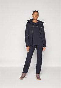 Tumši zila āra jaka ar rāvējslēdzēju, kapuci un logotipu, valkāta virs saskaņotas T-krekla un kombinēta ar tumšām biksēm un brūnām pārgājienu kurpēm.