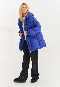 Modrá prošívaná péřová bunda s kapucí, vybavená prošívanou strukturou a několika kapsami. Zapínání na zip s růžovými detaily. Stylizováno s černými kalhotami.