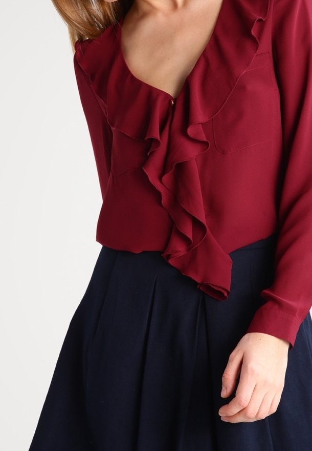 Donna che indossa una blusa lunga bordeaux con un davanti arricciato e una gonna plissettata blu navy, con la mano poggiata sul fianco.
