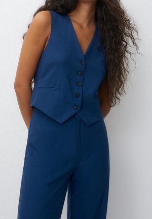 Vrouw draagt een mouwloos marineblauw vest met knopen, gecombineerd met bijpassende hoge taillebroek, staand tegen een effen witte achtergrond.