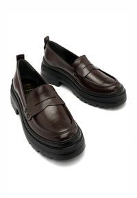 Brązowe skórzane buty wsuwane o niskim profilu, z przeszyciami i teksturowaną czarną gumową podeszwą. Posiadają akcentującą paskę z przodu.