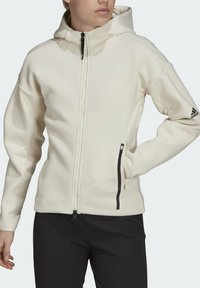 Bílá kapucínová bunda vyrobená z měkké látky s dlouhým zipem, bočními kapsami, černými akcenty a logem Adidas na rukávu.