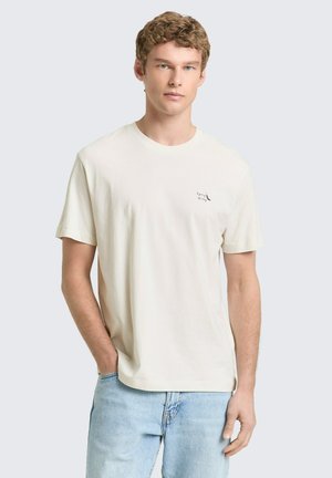 Jonge man met lichtbruin krullend haar, draag een eenvoudig off-white T-shirt met kleine zwarte tekst op de borst en lichtblauwe spijkerbroek.