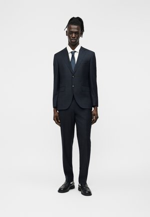 Homme noir debout, portant un costume bleu marine foncé à deux boutons, une chemise blanche, une cravate bleue marine et des chaussures en cuir noir, sur un fond blanc uni.