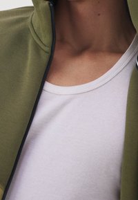 Primer plano del cuello y la parte superior del pecho con una camiseta de cuello redondo blanca y una chaqueta verde oliva sin abrochar.